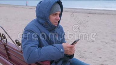 穿着蓝<strong>羽绒服</strong>坐在沙滩上的长凳上，在手机上的社交媒体上写一篇文章的人博主